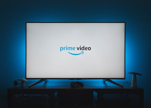 A television screen displays the Prime Video logo, situated on an entertainment unit in a dimly lit room. The room is illuminated with a subtle blue backlight, creating a calming atmosphere. Two small decorative items, resembling mushrooms, are placed on either side of the unit.