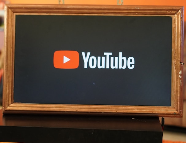 A rectangular screen encased in a wooden frame displaying the YouTube logo, characterized by a red play button icon next to the word 'YouTube' on a black background. The screen is placed on a small black table in a room with an orange wall and a couch in the background.